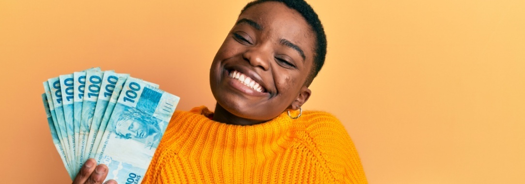 A photo of a young woman wearing a yellow knitted sweater with a big smile holding a wad of dollars against a yellow background